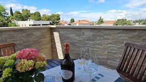 Una mesa con una botella de Prosecco, dos copas de vino y un jarrón de flores en una terraza con paredes de piedra y vistas a casas.