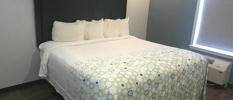 A neatly made bed with a white and green patterned bedspread, a dark headboard, and a window with blinds.