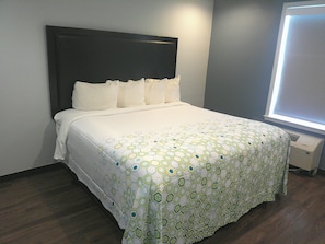 Una cama bien hecha con una colcha blanca y verde estampada, cabecera oscura y una ventana con persianas.