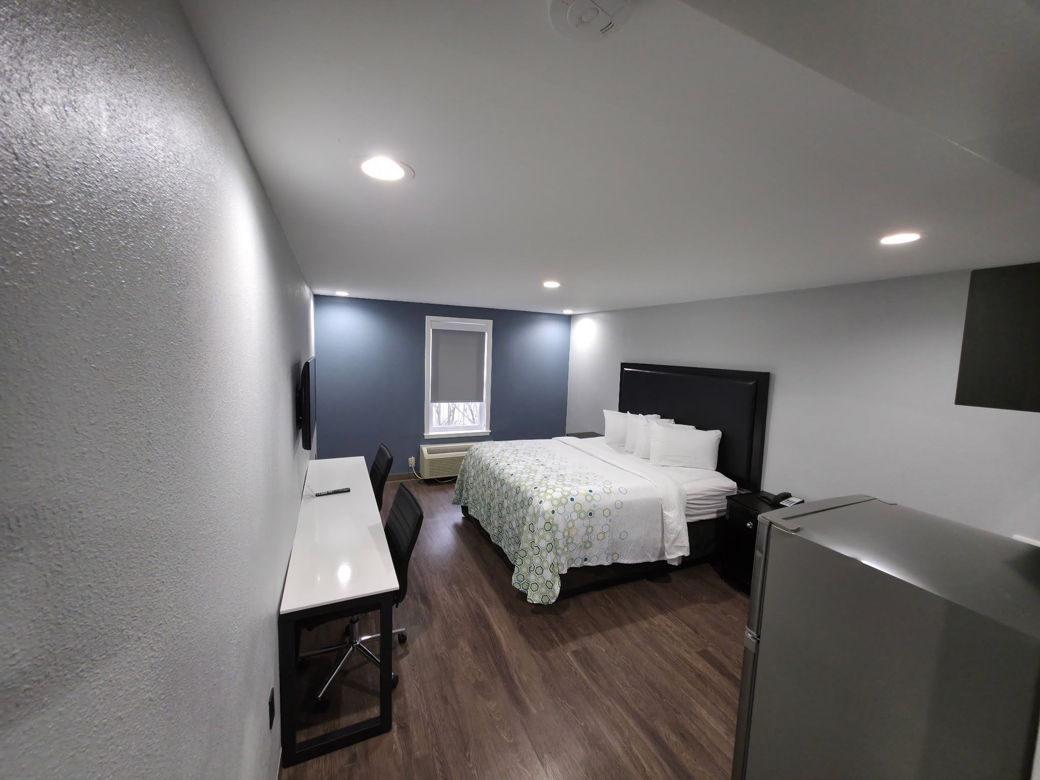 Ein Hotelzimmer mit einem Bett, einem Schreibtisch, einem Stuhl, einem kleinen Kühlschrank und einem Fenster mit Jalousien.