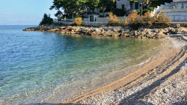 Une zone côtière avec une plage de galets, une eau claire et un bâtiment aux murs de pierre, entouré d’arbres.