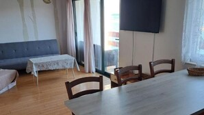 Un coin repas avec une table et des chaises, un canapé, une petite table recouverte d’une nappe à motifs et un téléviseur fixé au mur.