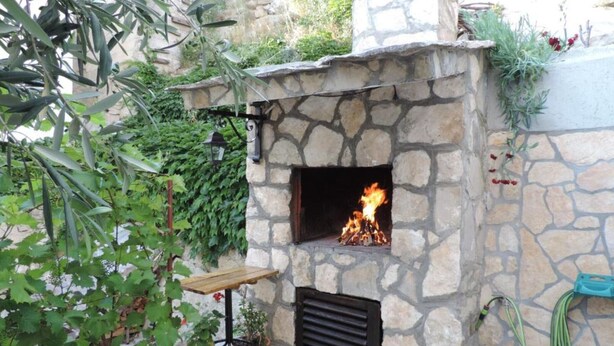 A stone-built outdoor fireplace with a fire burning inside, surrounded by potted plants and a small table. - Apartments Rubić - Onebedroom Apartment with Loggia and SeaView (2) (Omis)