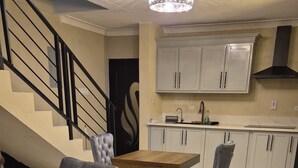 Ein Wohnzimmer mit einem grauen Ecksofa, zwei gelben Kissen, einem gemusterten Teppich, einer Treppe und einer Küchenzeile mit weißen Schränken und einer schwarzen Dunstabzugshaube.
