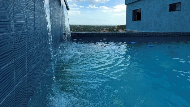 Ein Schwimmbecken mit Wasserfall, ein klarer Himmel und ein Gebäude im Hintergrund.
