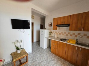 A compact kitchen with wooden cabinets, a white refrigerator, a small countertop with a sink, and a television mounted on the wall. - Apartments Rubić - TwoBedroom Apartment with Loggia and SeaView (4) (Omis)