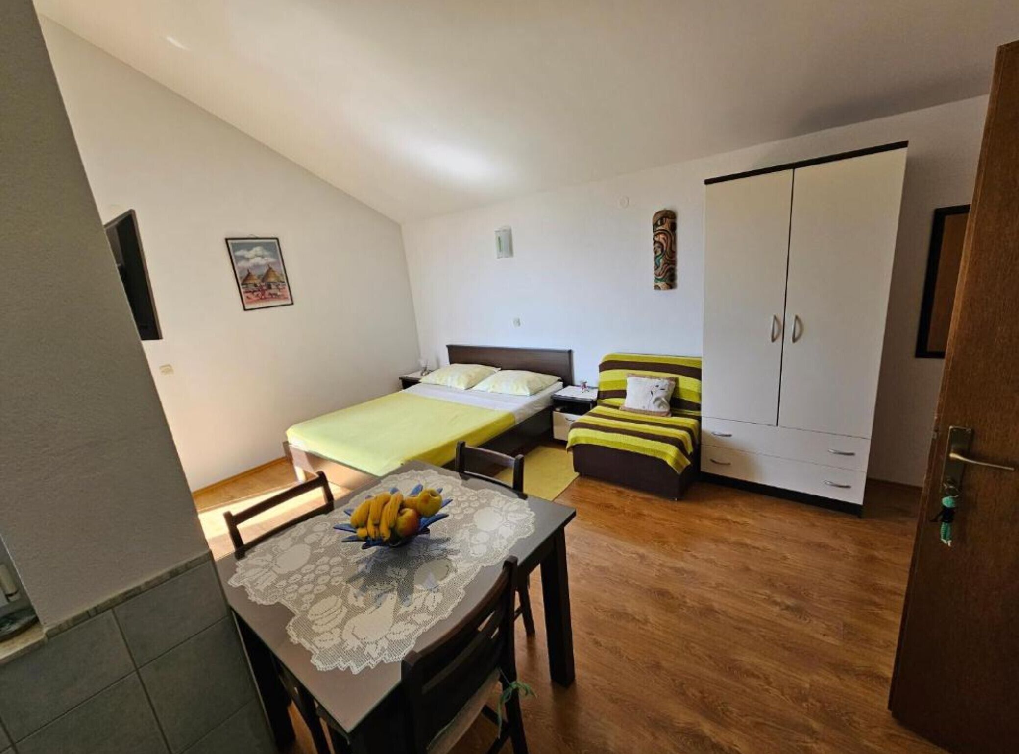 A small, well-lit room with a bed, a sofa, a table with fruit, and a wardrobe.
