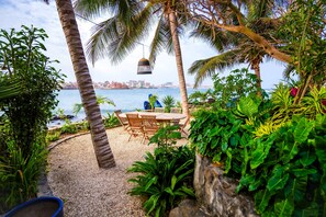 A patio with a table and chairs, surrounded by palm trees and plants, overlooking a body of water and a distant cityscape. - Villa Ipomée and its waterfront tropical garden on Ngor Island (Dakar)