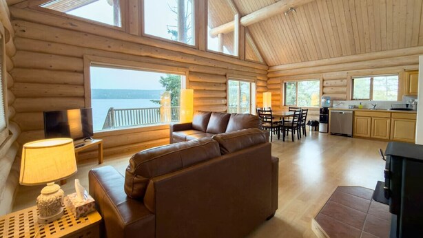 A log cabin with a living area featuring a sofa, a TV, and a dining table with chairs. There is a kitchen area with a stove and cabinets. Large windows provide views of the outdoors. - Serene Lakeside Escape – Juniper Lodge, Lac La Hache (Lac la Hache)