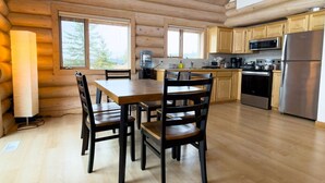 A log cabin interior with a wooden dining table and chairs, a kitchen with modern appliances, and large windows offering a view of the outdoors. - Serene Lakeside Escape – Juniper Lodge, Lac La Hache (Lac la Hache)