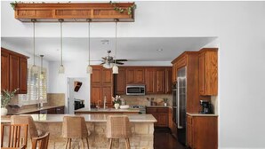 Eine moderne Küche mit Holzschränken, einer zentralen Kochinsel mit Barhocker, einem Deckenventilator und Edelstahlgeräten.