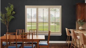 Ein Essbereich mit Tisch und Stühlen, ein Fenster mit Jalousien, ein Blumentopf und ein Küchenbereich mit einem Schrank und einer Vase.