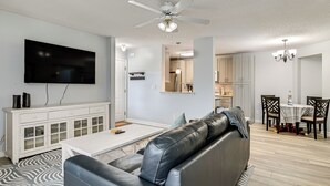 Uma sala de estar moderna com um sofá de couro, uma televisão de tela plana, uma área de jantar com mesa e cadeiras, e uma cozinha ao fundo.