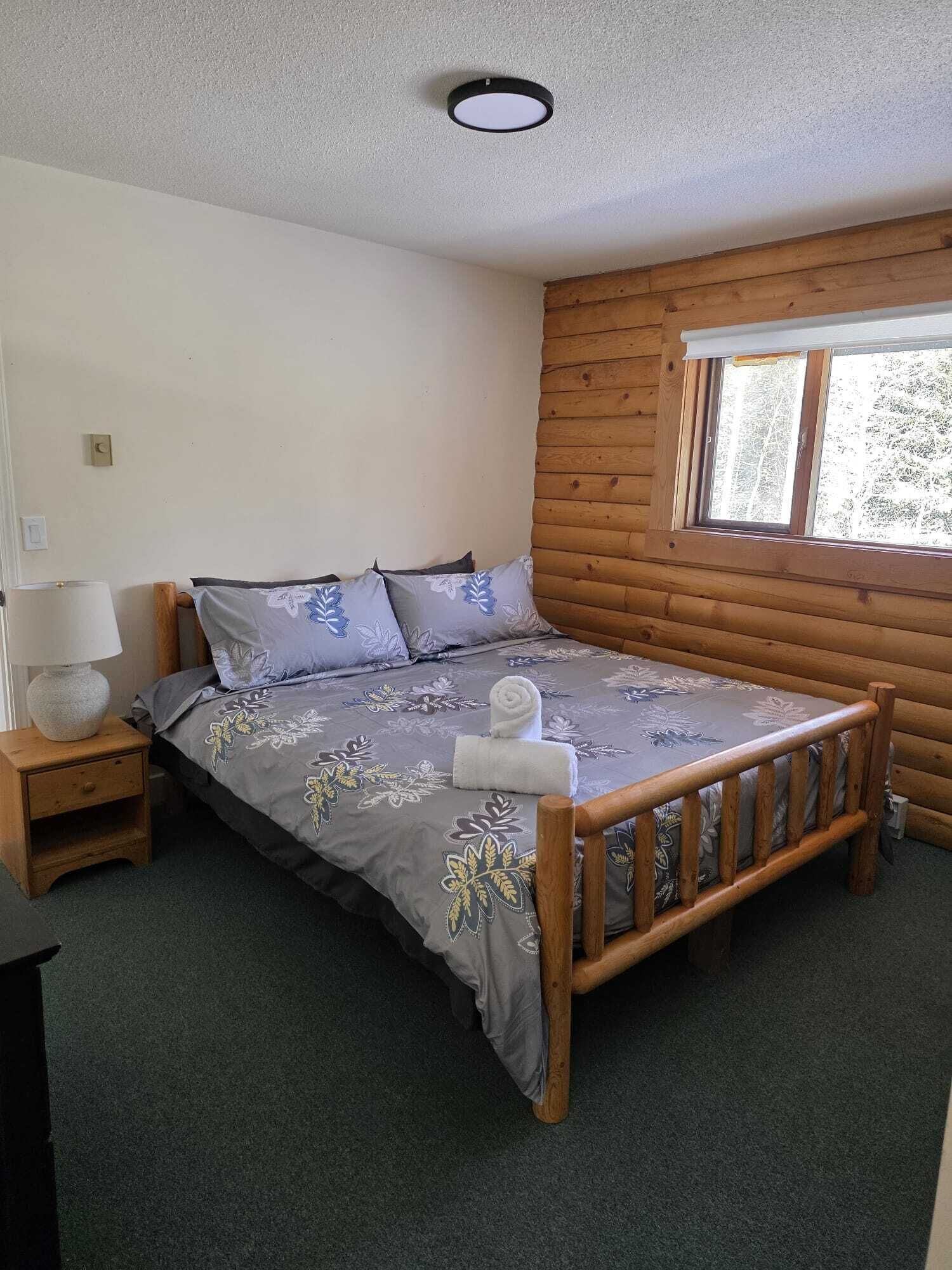 Un letto singolo con struttura in legno e copripiumino floreale grigio, un comodino con una lampada e una finestra vista alberi.