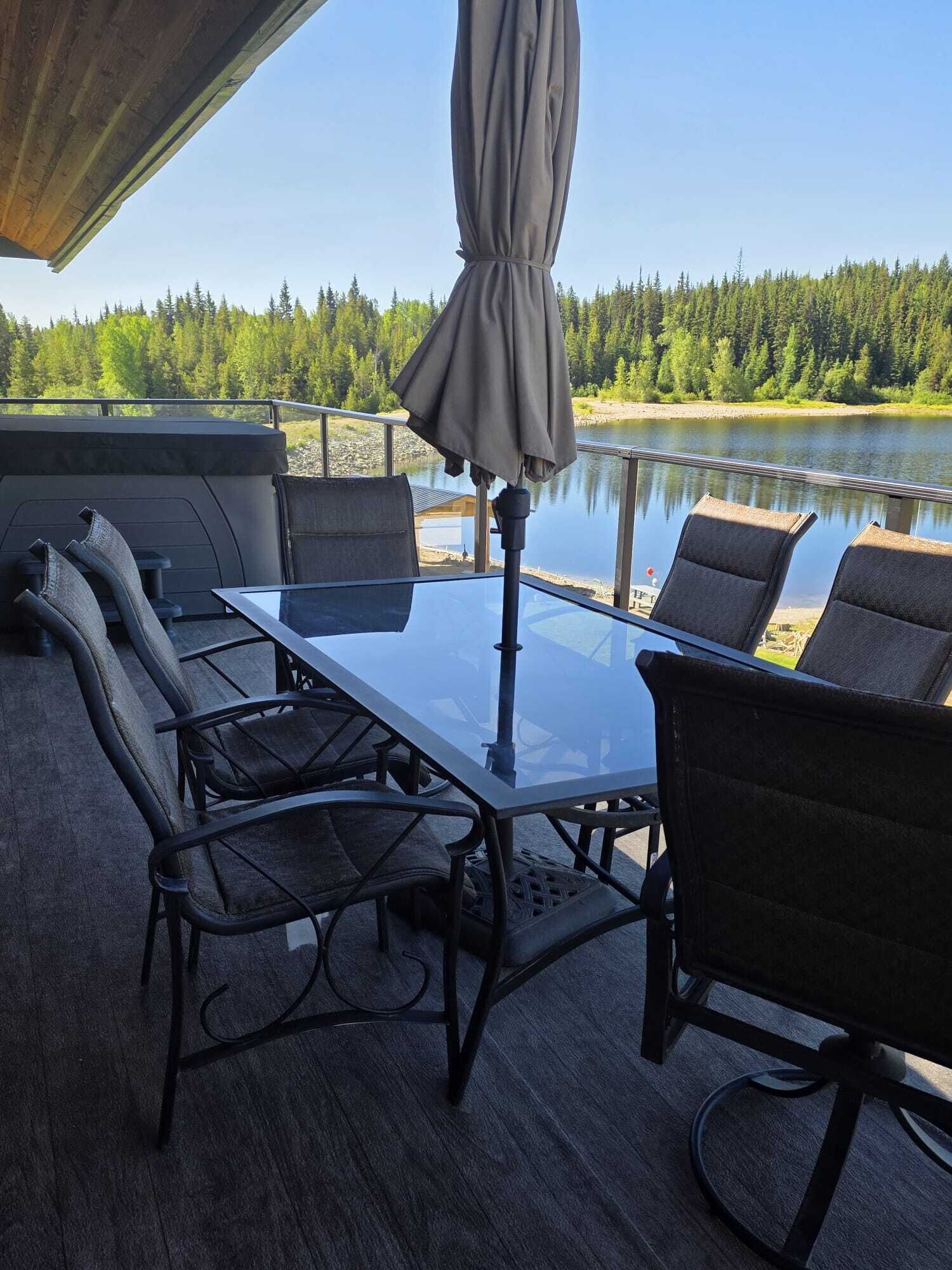 En utomhusplats med sittmöbler, bestående av ett glasbord och stolar, ett parasoll och utsikt över en sjö och en skog.