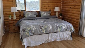 A bedroom with a wooden log wall, a large bed with floral bedding, two bedside lamps, and a window with a forest view. - 4 Bdrm Lodge Suite w Hot Tub, beautiful Lake views! Pet Friendly (Naramata)