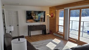 A modern living room with a large TV, a sofa, a coffee table, and a view of a snowy outdoor area through glass doors. - 4 Bdrm Lodge Suite w Hot Tub, beautiful Lake views! Pet Friendly (Naramata)