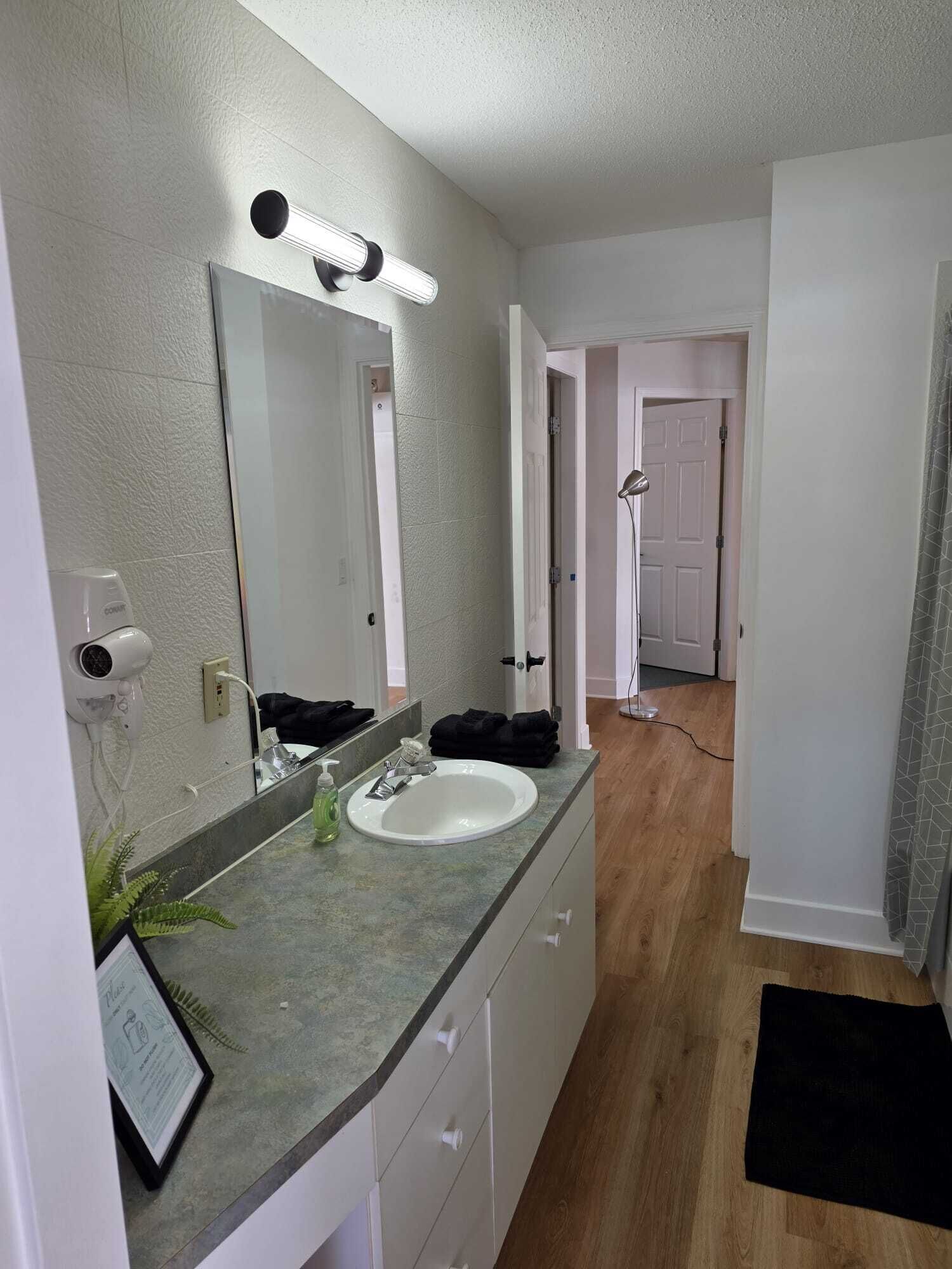 A bathroom with a white sink, a mirror, and a hairdryer on the wall. There is a black towel on the counter.