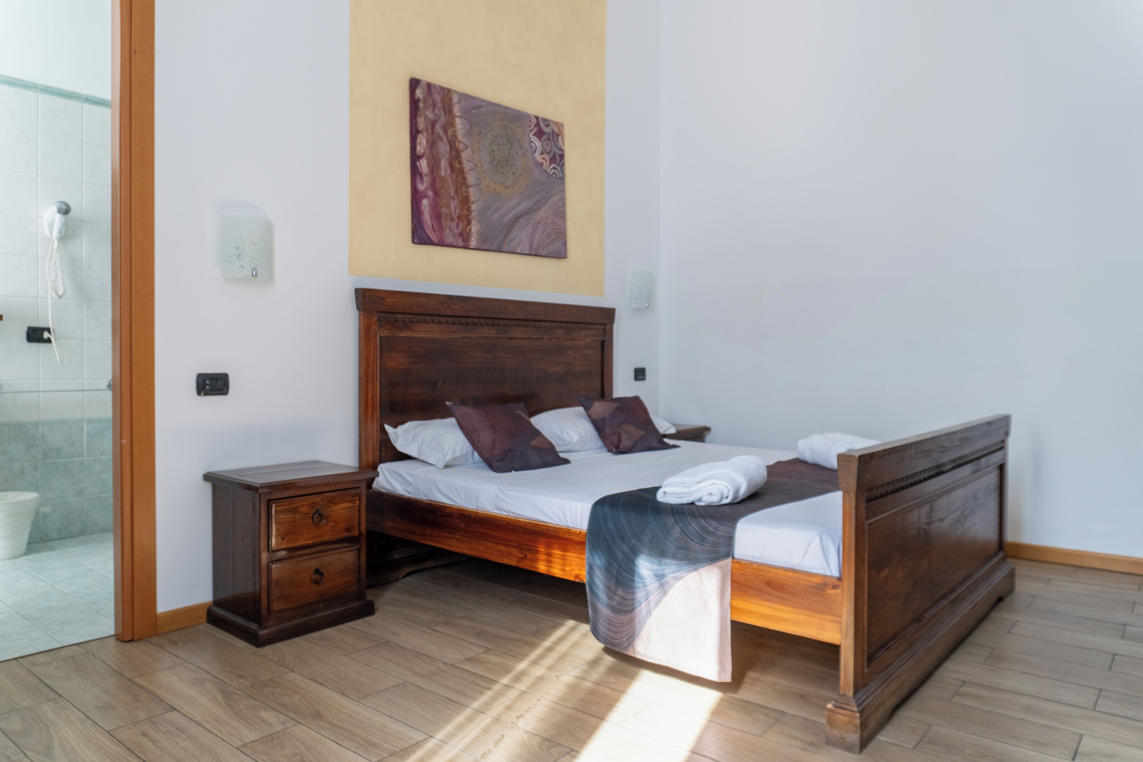 A bedroom with a wooden bed, a nightstand, a bathroom with a shower, and a wall-mounted artwork.