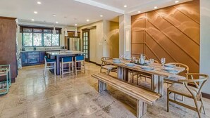 A spacious dining area with a long wooden table set for six, a kitchen in the background, and a modern interior design. - Modern Condo + Pool Access | Gated Community (Coco)