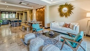 A spacious living area with a sofa, coffee table, and chairs. A kitchen is visible in the background. - Modern Condo + Pool Access | Gated Community (Coco)