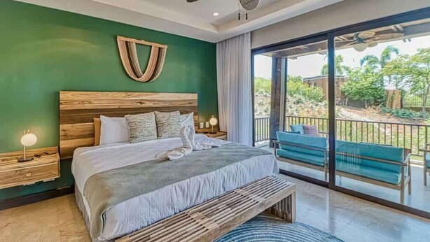 A bedroom with a wooden headboard, a large bed with white linens, a bedside table with a lamp, and a view of a balcony with outdoor seating. - Modern Condo + Pool Access | Gated Community (Coco)