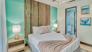 A bedroom with a bed, two bedside tables, a wall-mounted lamp, a wooden slat partition, and artwork on the wall. - Modern Condo + Pool Access | Gated Community (Coco)