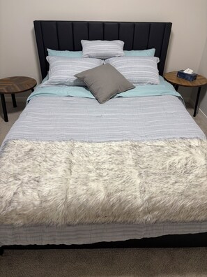 Een bed met een gestreept dekbed, een grijze kussen en een zachte witte deken. Er staan twee houten nachtkastjes, waarvan er op één een tissuedoosje staat.