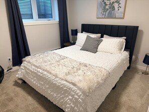 Un dormitorio con una cama grande, cabecero, dos lámparas de noche, una ventana con cortinas y una obra de arte colgada en la pared.