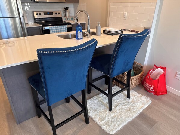 A kitchen with a central island, two blue upholstered chairs, a stainless steel refrigerator, a black stove, and a white tiled backsplash.