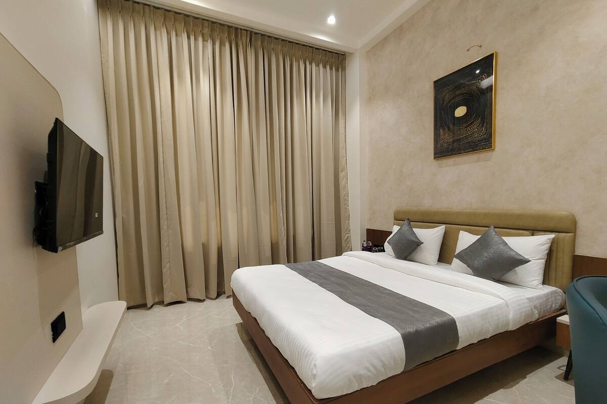 A hotel room with a large bed, a flat-screen TV mounted on the wall, and a window with beige curtains.