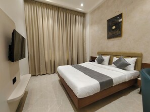 Una habitación de hotel con una cama grande, un televisor de pantalla plana montado en la pared y una ventana con cortinas color beige.