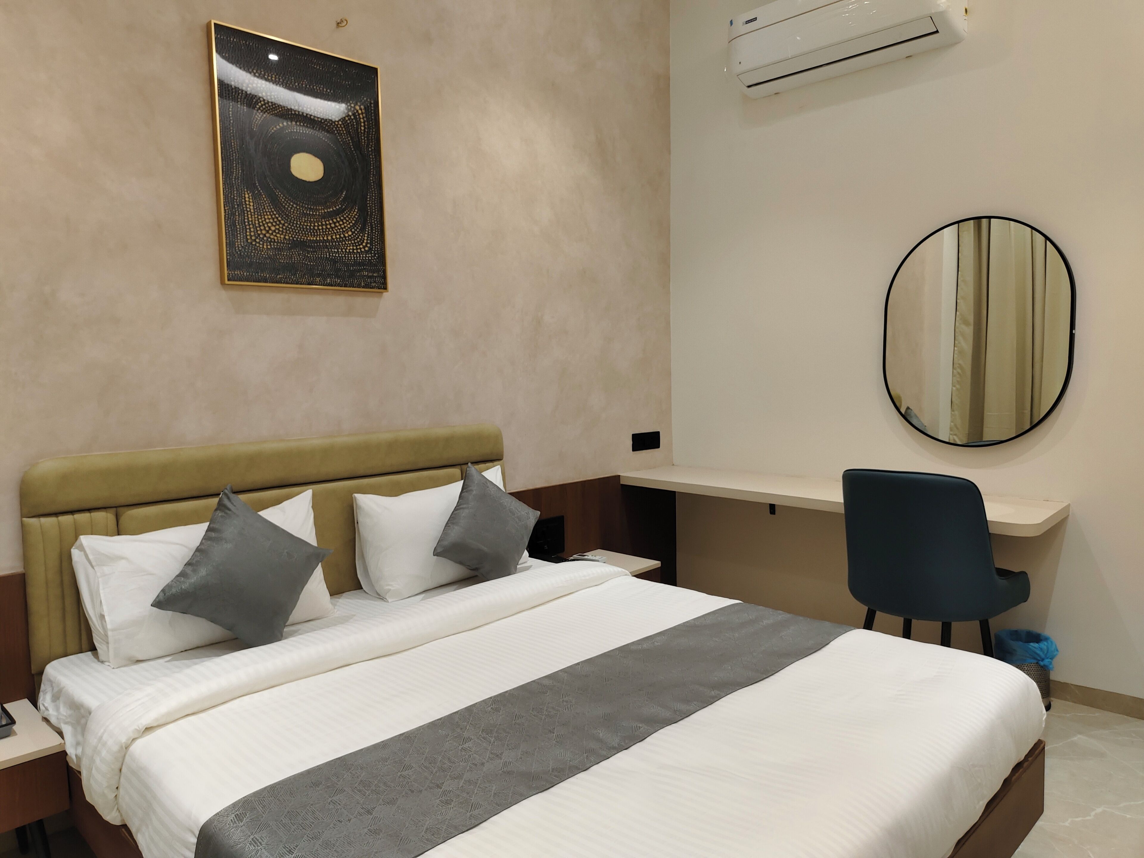 A hotel room with a bed, a desk, a chair, an air conditioner, and a wall-mounted mirror.