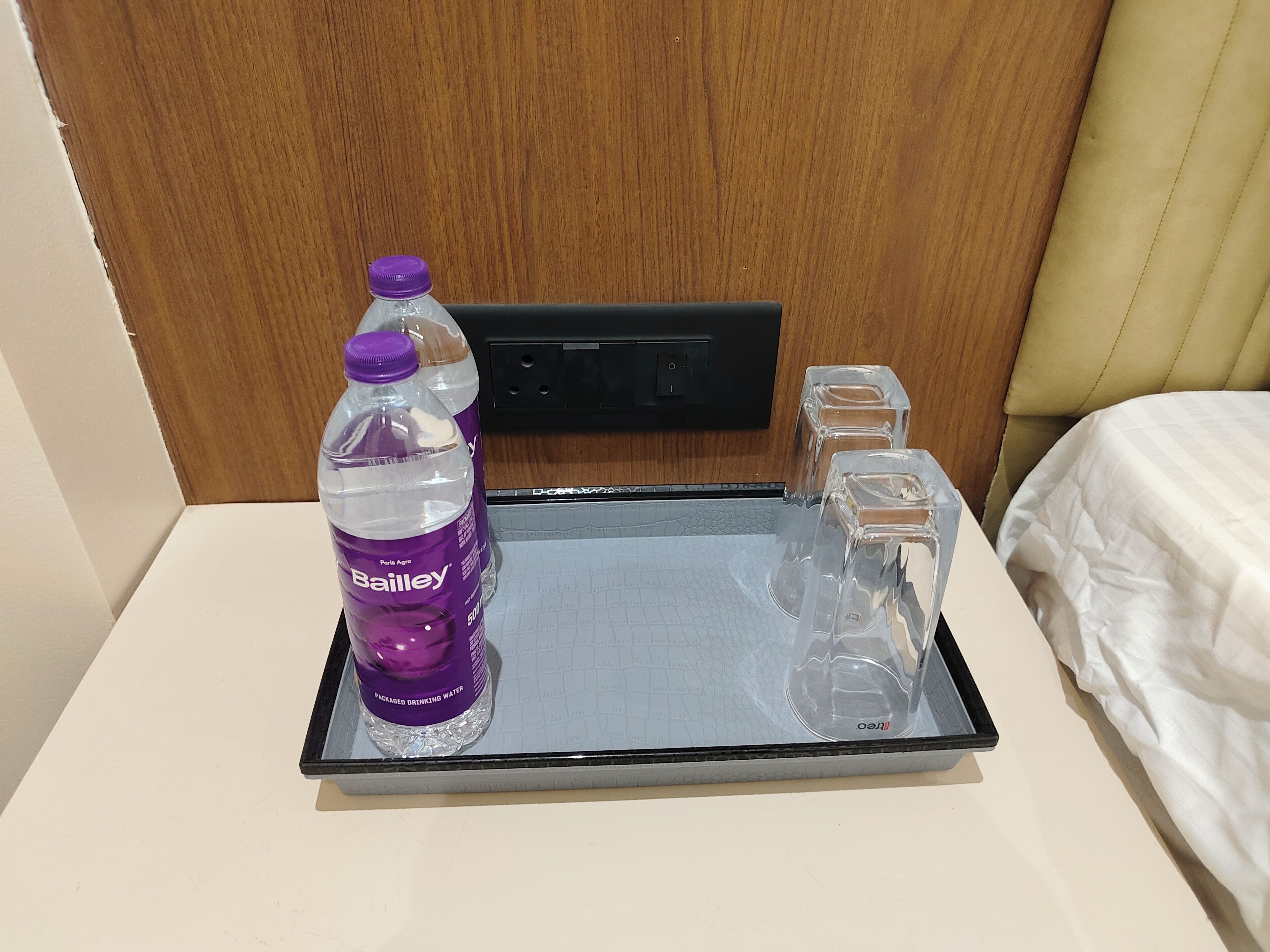 A hotel room with a bed, a nightstand, a tray with a bottle of Bailey's water, and two glasses.