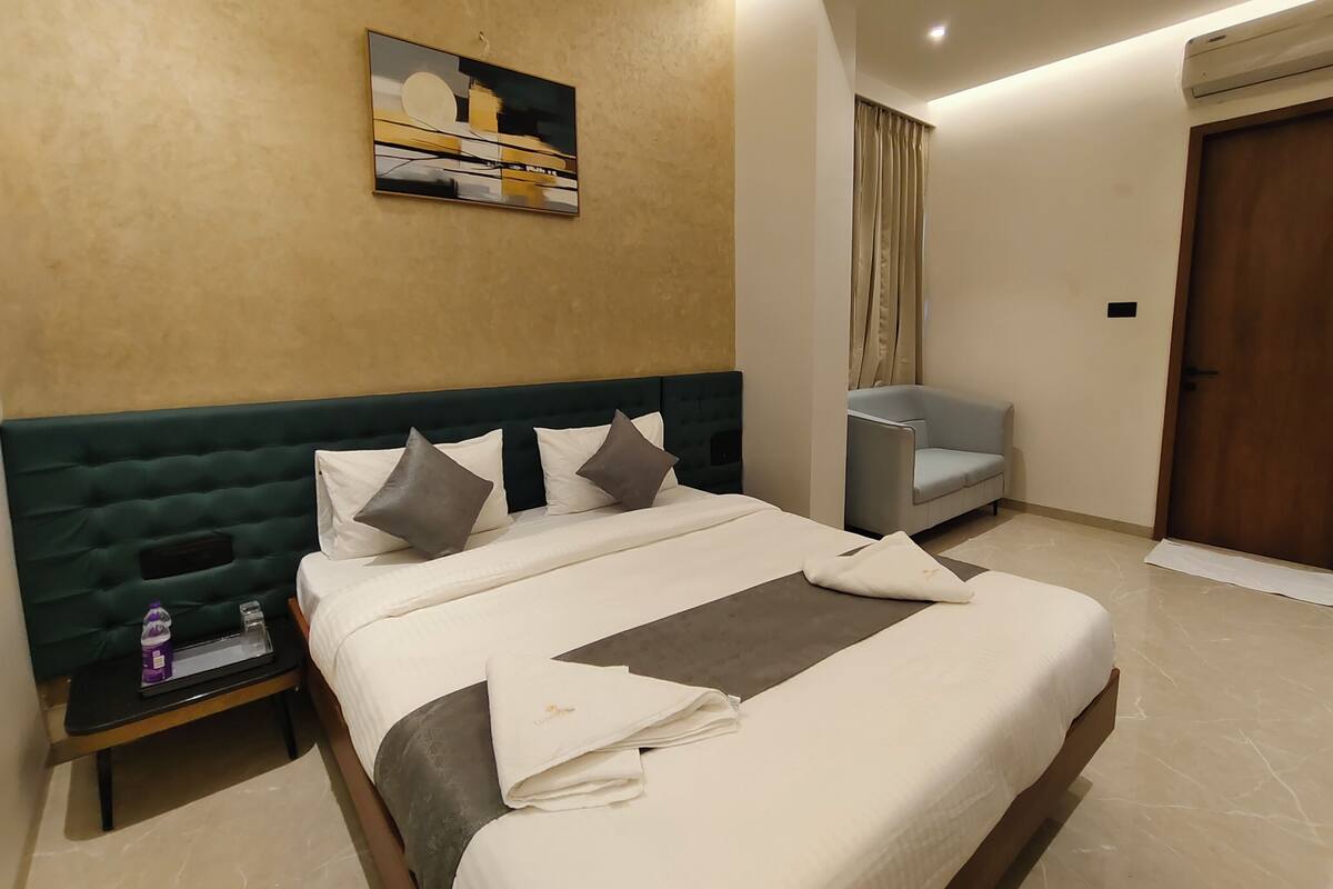 A hotel room with a large bed, a small table with a bottle and glass, a grey sofa, and a door.