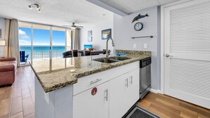 A modern kitchen with a granite countertop, a sink, and a dishwasher. There is a dining area with a table and chairs, and a view of the ocean through large windows. - Long Beach Resort 4-605 (Panama City Beach)
