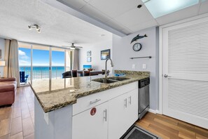 A modern kitchen with a granite countertop, a sink, and a dishwasher. There is a dining area with a table and chairs, and a view of the ocean through large windows. - Long Beach Resort 4-605 (Panama City Beach)