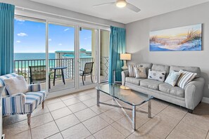 A living room with a glass coffee table, a sofa, and a chair, a balcony with a view of the ocean, and a painting on the wall. - Ariel Dunes I 1306 (Miramar Beach)