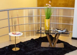 A modern room with a glass table, a small round table with books, a black furry rug, and a yellow vase with flowers.