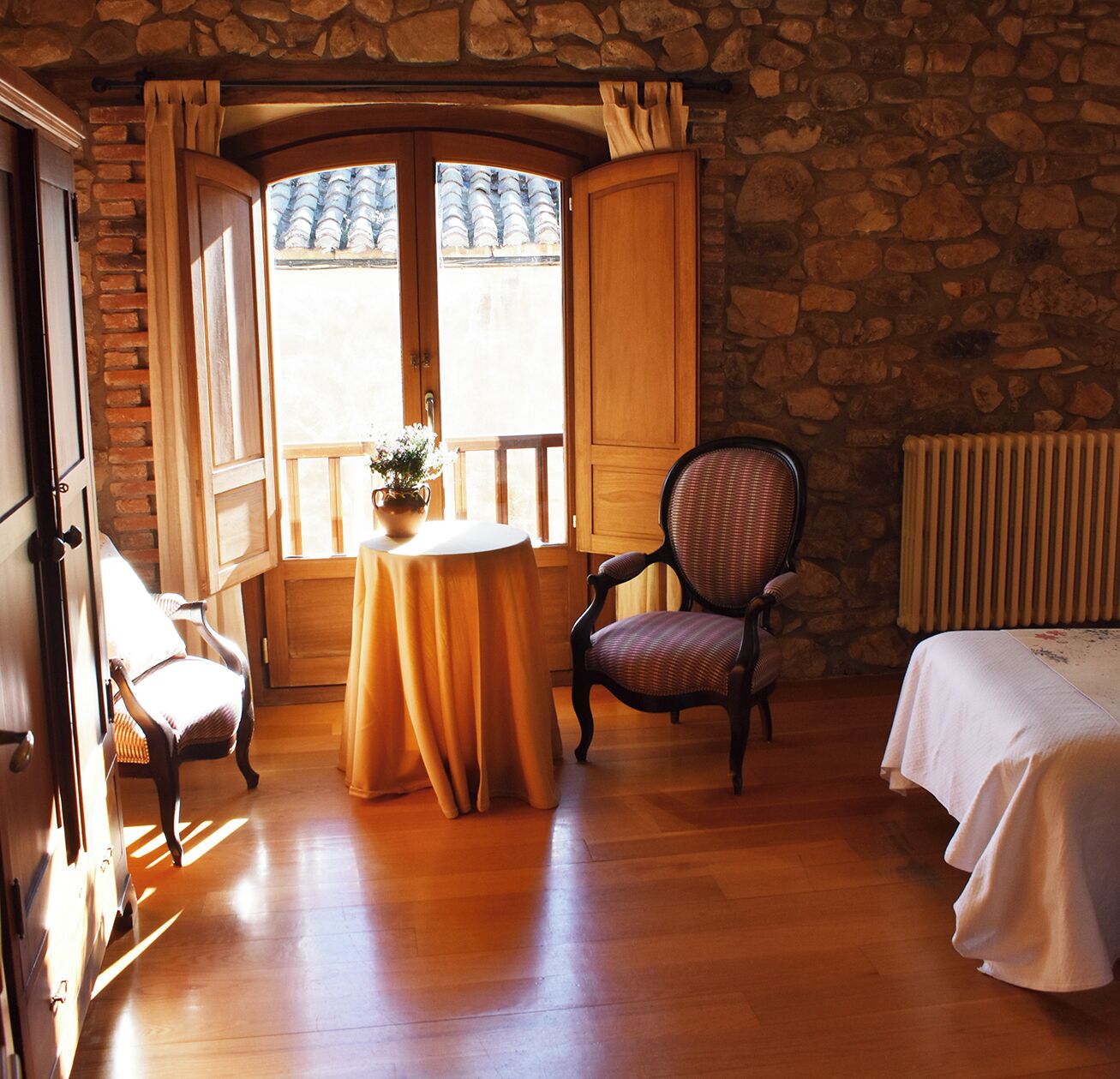 A room with a stone wall, a wooden table with a vase, a wicker chair, and a bed with a white bedspread.