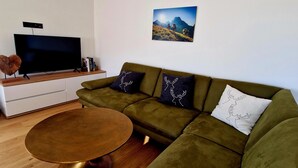 Een moderne woonkamer met een groene bank, een ronde salontafel, een flatscreen televisie en een schilderij van een berglandschap aan de muur.