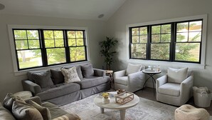 A living room with a grey sectional sofa, a white armchair, and a round coffee table. There are large windows with a view of greenery outside. - North Fork Waterfront Oasis in Mattituck (Mattituck)