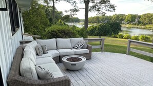 A covered outdoor deck with a sectional sofa, a fire pit, and a view of a lake and greenery. - North Fork Waterfront Oasis in Mattituck (Mattituck)
