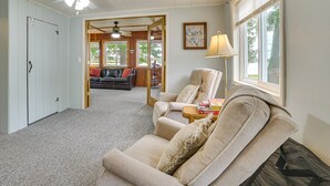 Ein Wohnzimmer mit einem beigefarbenen Sofa, einem Holztisch und einem Deckenventilator. Eine Tür führt in einen anderen Raum, ein Fenster mit Jalousien ist ebenfalls vorhanden.