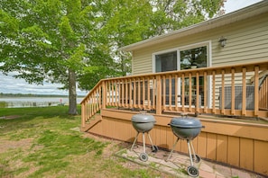 A house with a wooden deck, two barbecue grills, and a view of a lake.