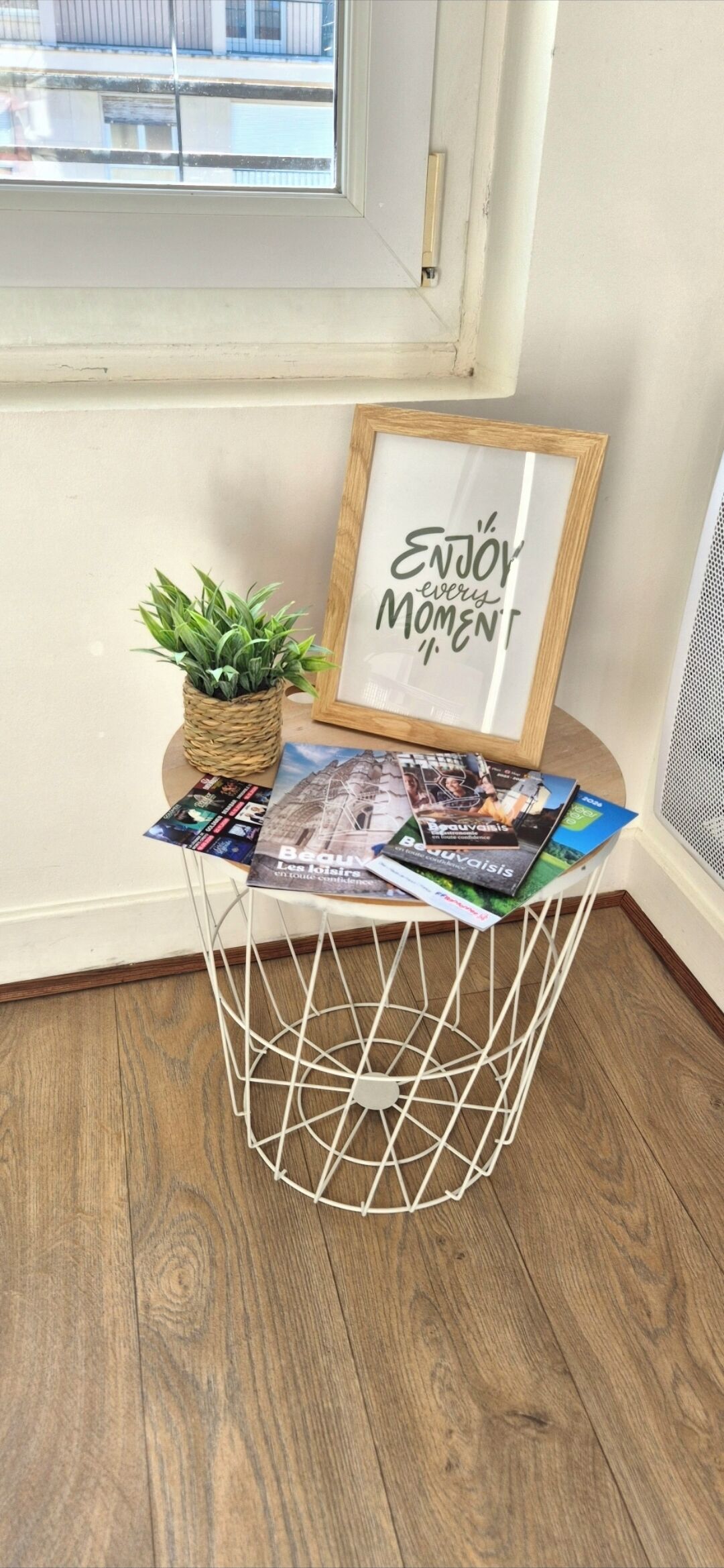 Une petite table en bois avec un socle en fil métallique blanc, un pot à plantes en osier et un cadre contenant une citation inspirante.