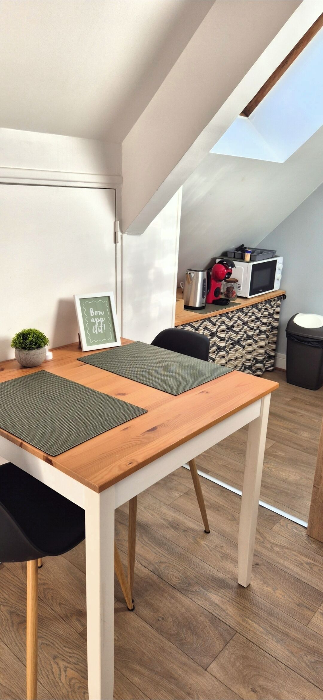 Une petite pièce agréable et chaleureuse, avec une table en bois, deux tapis verts, un panneau encadré, une petite plante et un coin cuisine équipé d’un four à micro-ondes et d’une bouilloire.