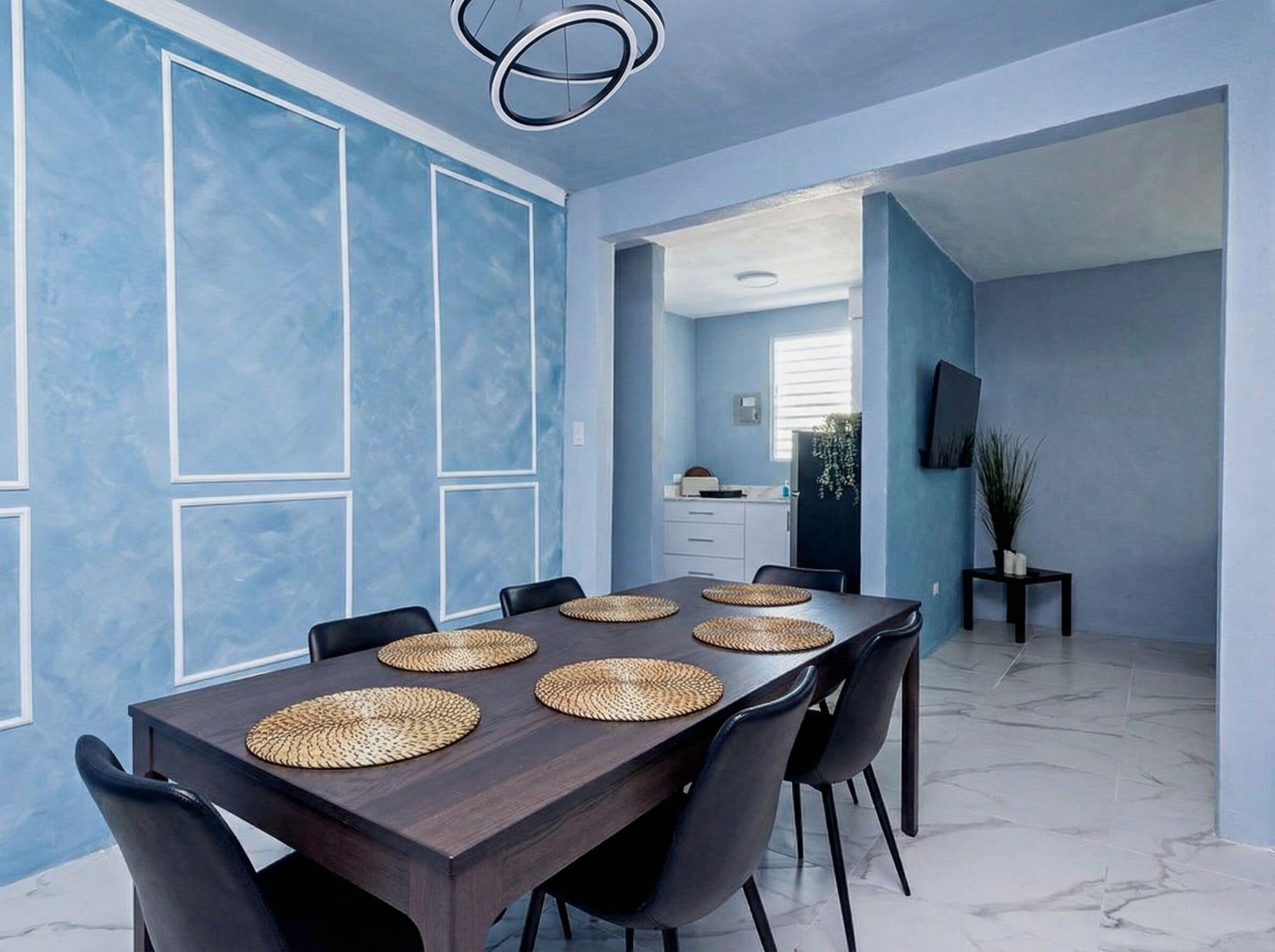 Un comedor con una mesa larga, sillas negras, individuales tejidas y una cocina al fondo.