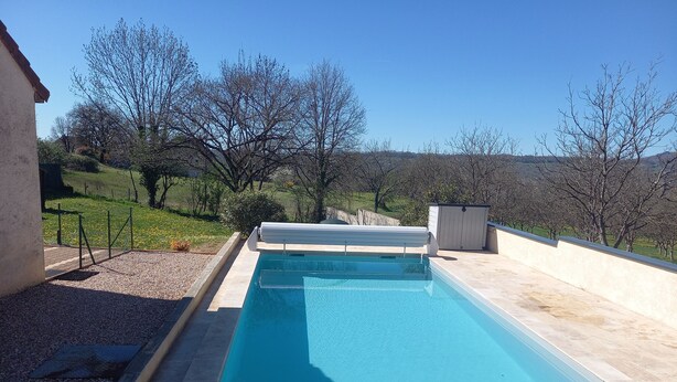 A rectangular swimming pool with a tiled deck, surrounded by a low wall and a grassy area with trees.