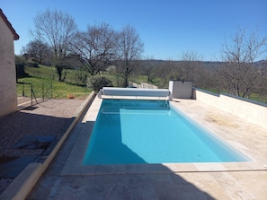 Une piscine rectangulaire avec une terrasse carrelée, entourée d’un petit muret et d’une zone herbeuse agrémentée d’arbres.
